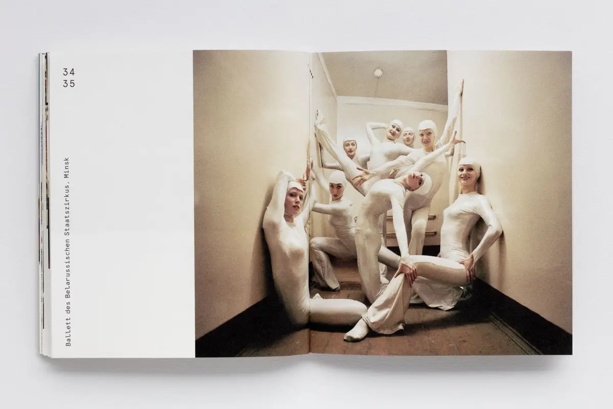 Group of ballet dancers in white unitards and caps posing on a staircase inside a beige studio.