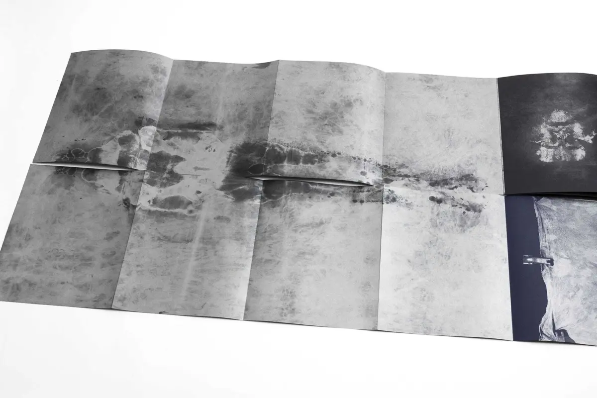 Gray-scale accordion-folded poster on a white surface with abstract ink-splatter textures across panels.