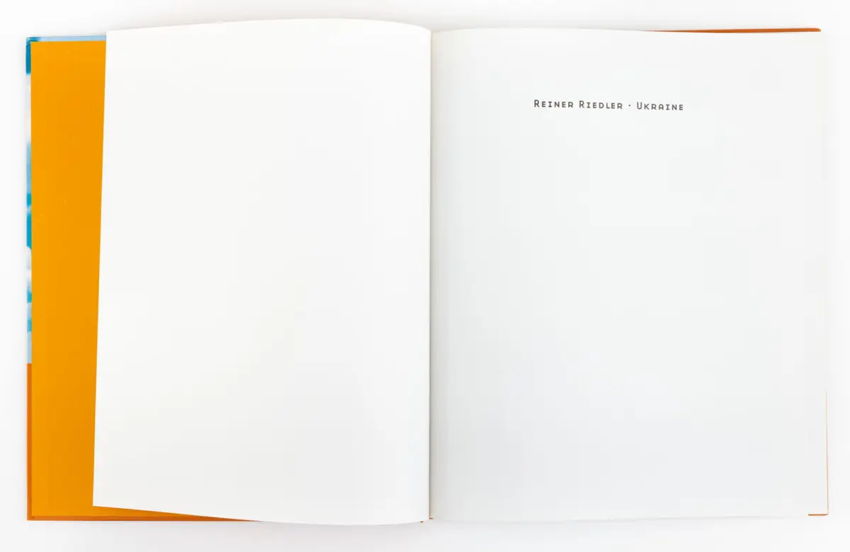 Open book with white pages; orange cover visible at left, right page displays REINER RIEDLER • UKRAINE.