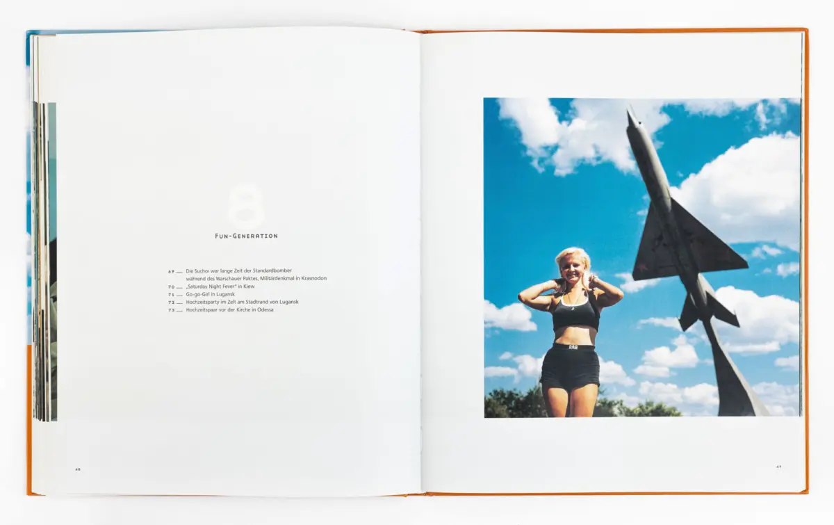 Athletic woman in sports bra and shorts posing with hands on head before a rocket monument outdoors under a blue sky.