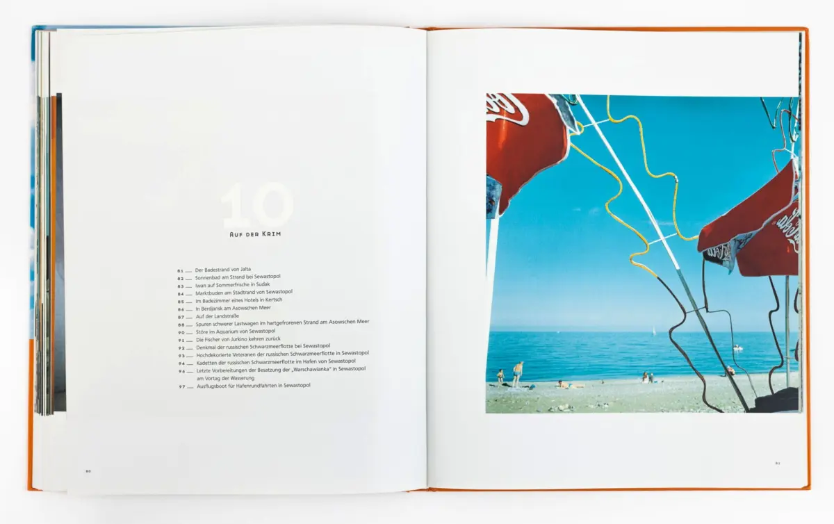 Open book spread; right page shows a bright beach scene with blue sea, red umbrellas, and people under a clear sky.