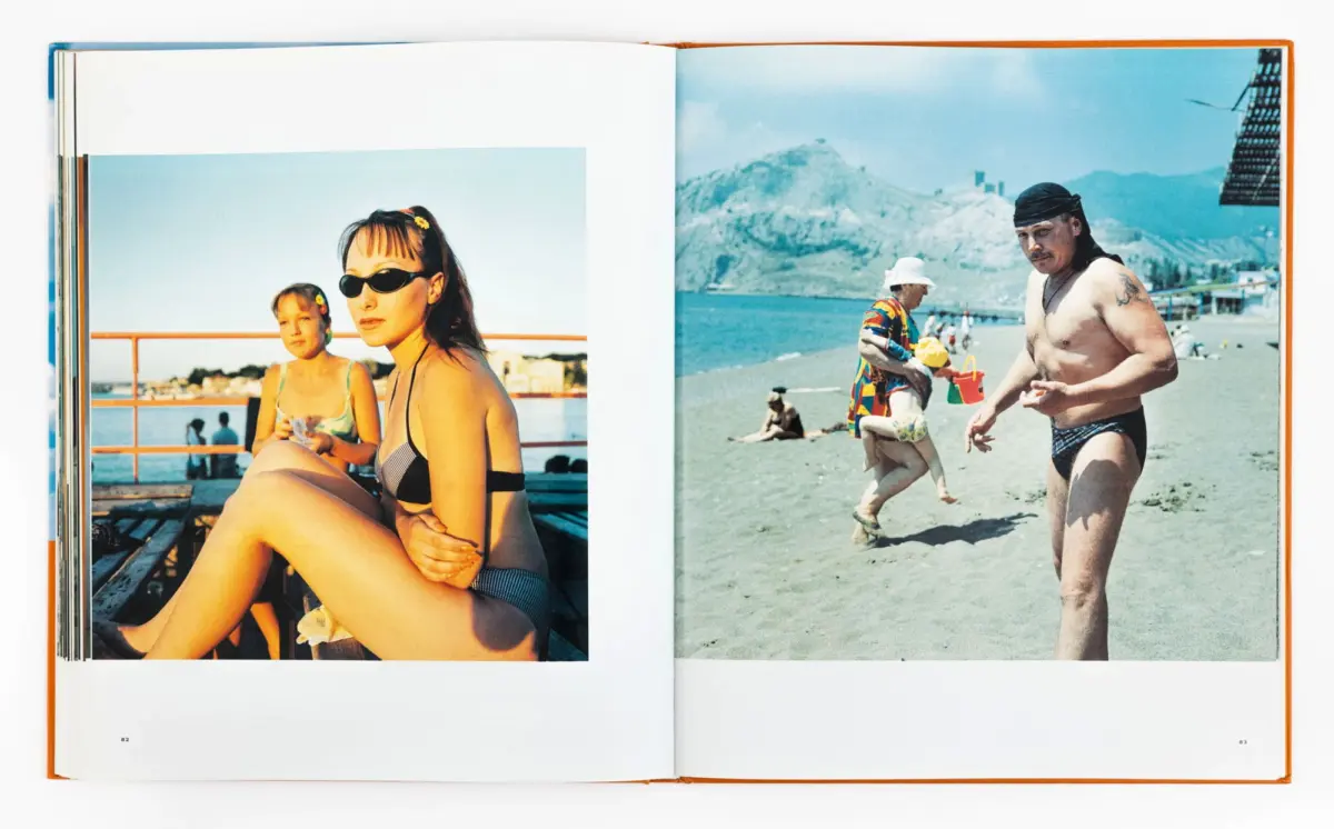 Two people in swimsuits sit on a seaside deck; on the right, a person in swimwear walks the sandy beach with mountains behind.