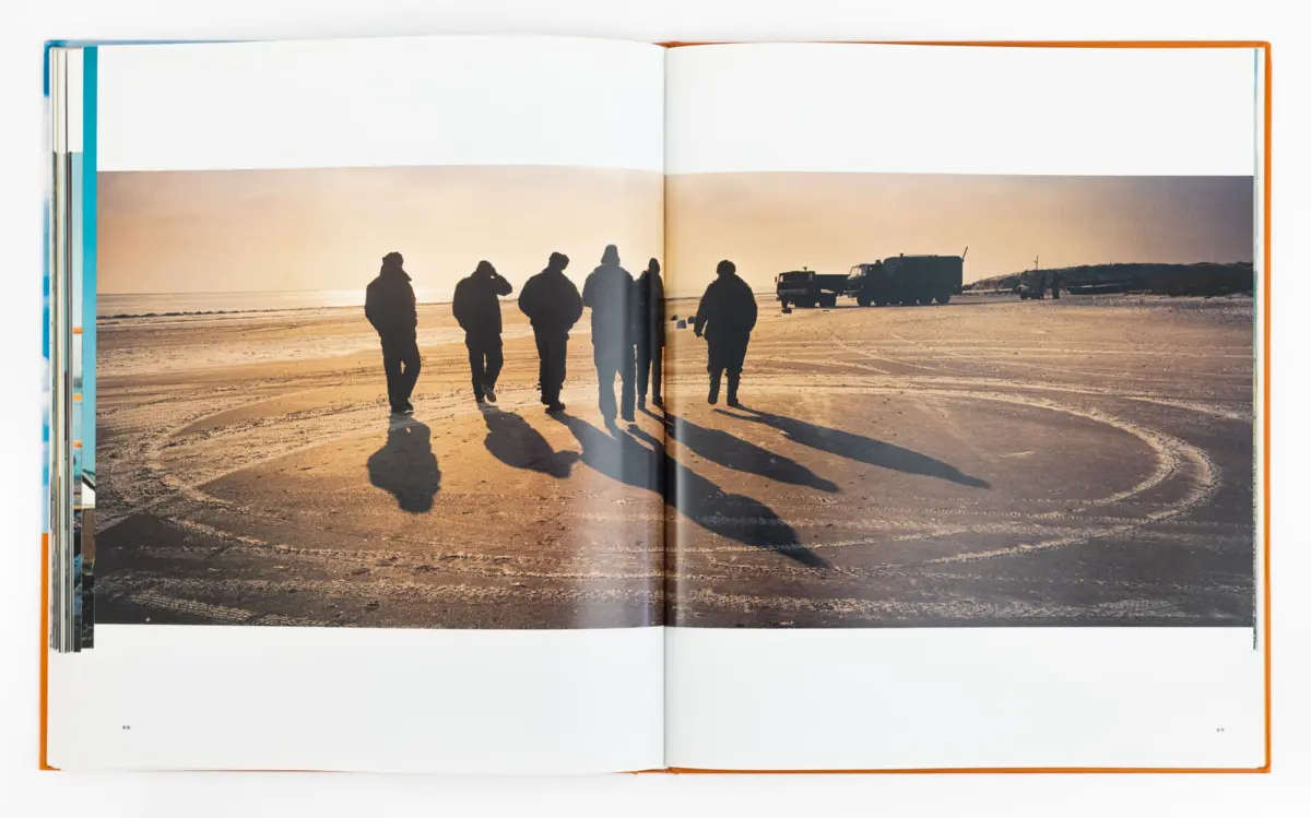 Silhouetted group of people standing on a sandy beach at sunset; long shadows stretch across wet sand with trucks in the distance.