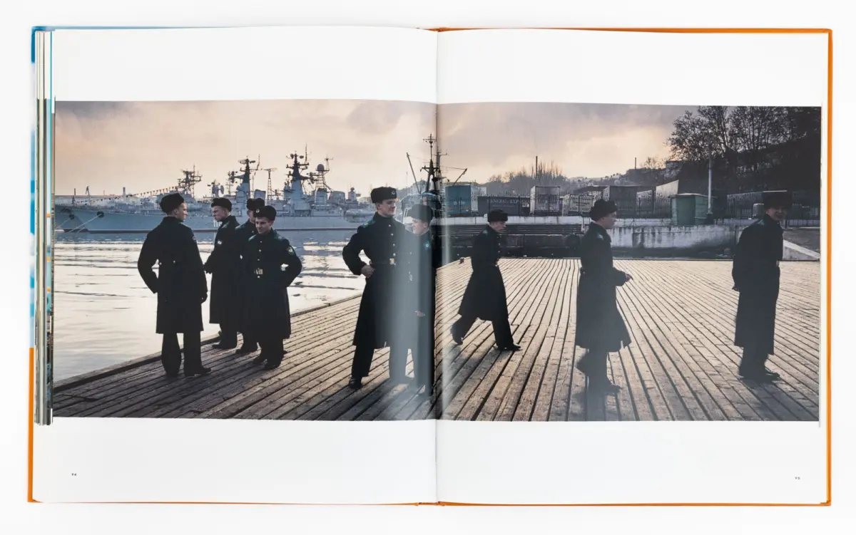 Naval sailors stand on a wooden pier at a harbor, with large warships and industrial buildings in the background.