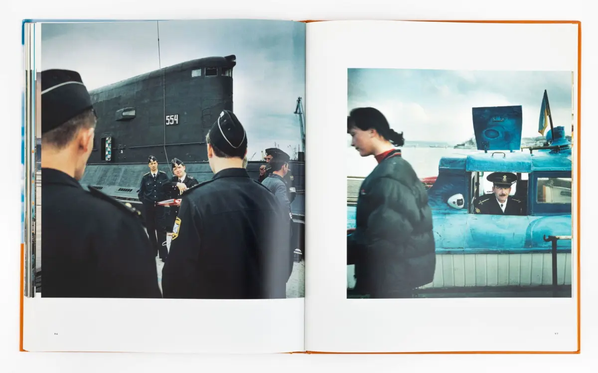 Naval officers in uniform stand on a dock beside a large submarine; on the right, a blue vehicle with a driver.