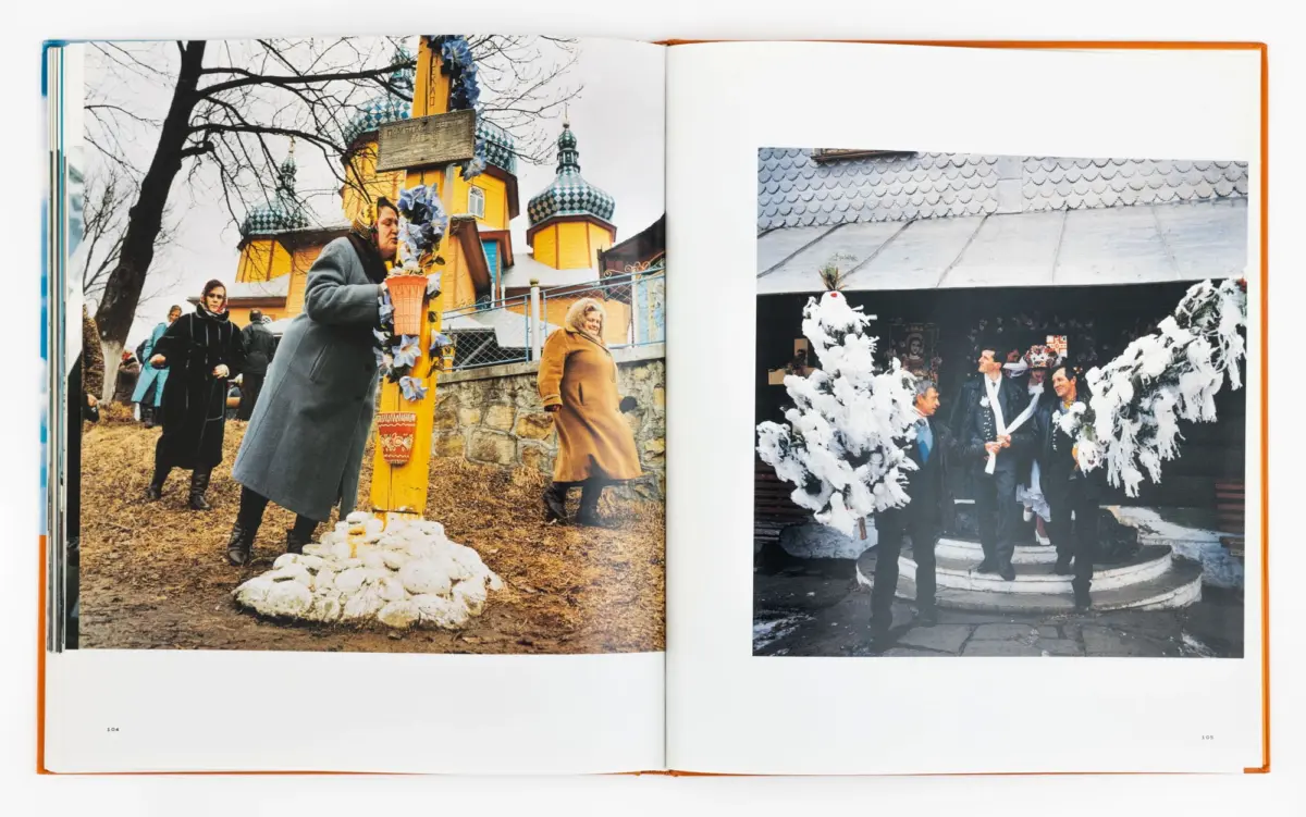Open book spread: gray-coated person leans toward a flower-decorated yellow cross beside an Orthodox church; snow on the ground; right page shows a formal gathering with white feather decorations.