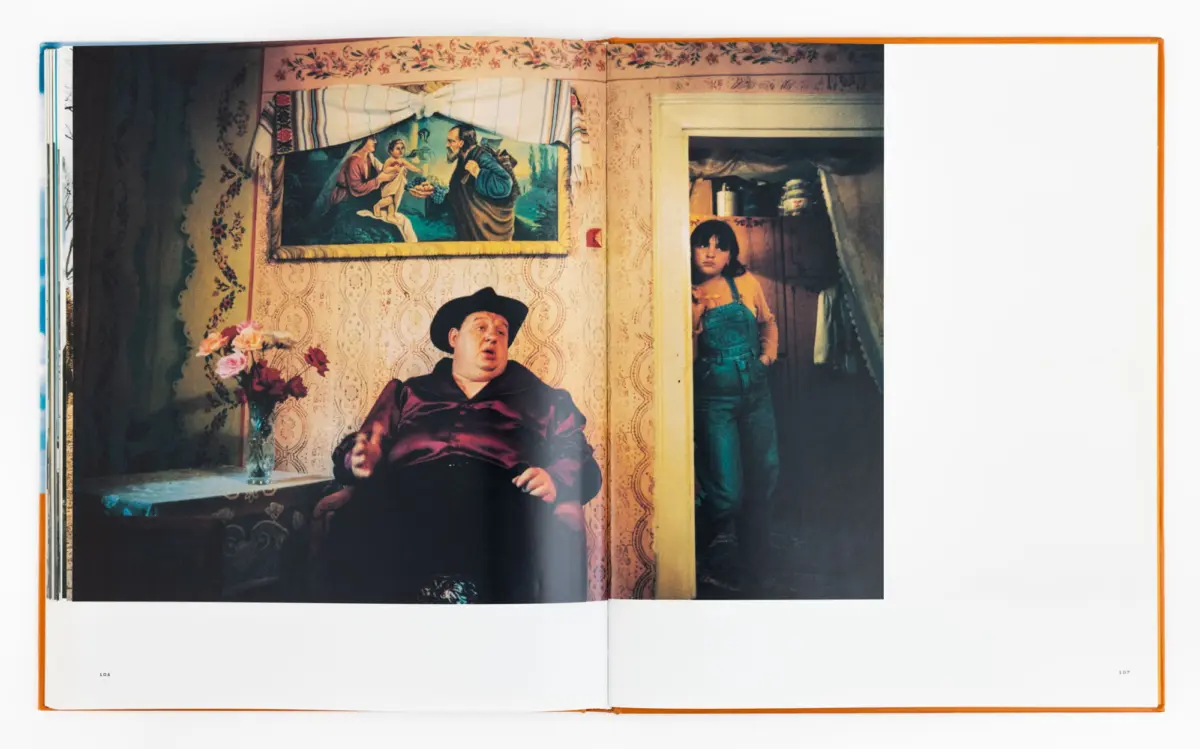 Portly man in a hat seated in a vintage living room; girl in denim overalls stands in doorway beside floral wallpaper and wall painting.