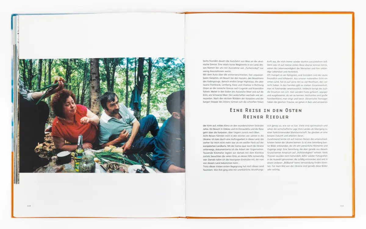 Group of people picnicking in a forest, sitting around a blanket with bottles and dishes amid green trees.