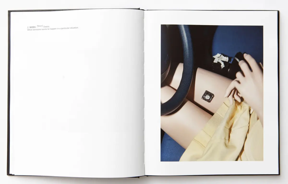 Open book page showing a photo of a person in a car, reaching toward a small square control on the dashboard near the steering wheel.