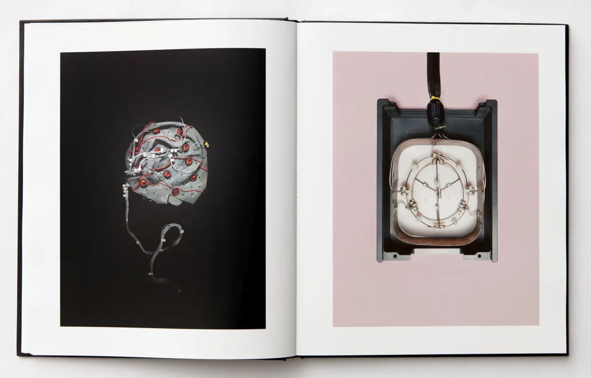 Open book showing left: a metallic brain sculpture wrapped in wires on black; right: a square medical implant mounted in a frame on pink.