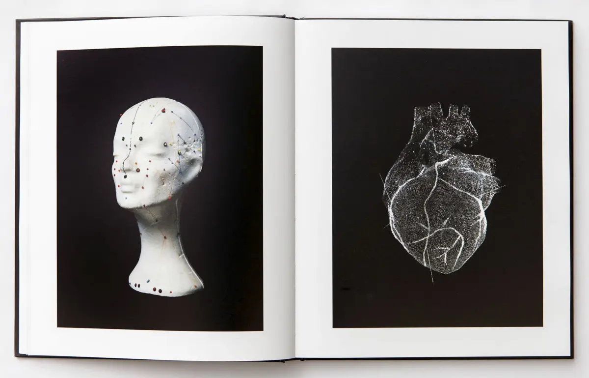 Open book displaying two images: left white mannequin head studded with colorful pins; right white line drawing of a heart on black background.