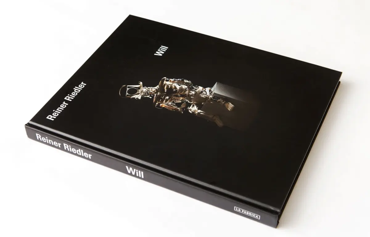 Closed black hardcover book on a white surface; cover shows a metallic humanoid robot in a space suit.