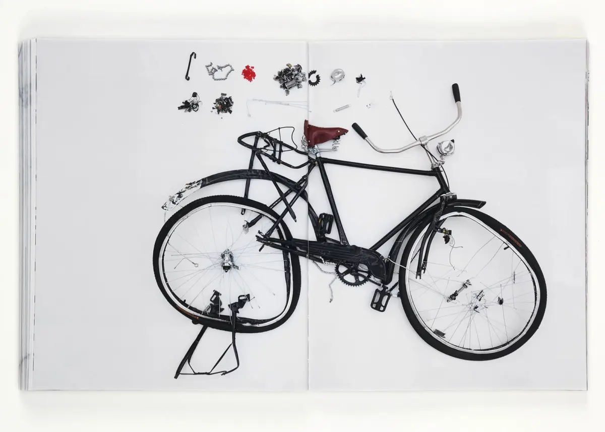 Black bicycle with red saddle and scattered spare parts arranged on white book pages in a flat lay.