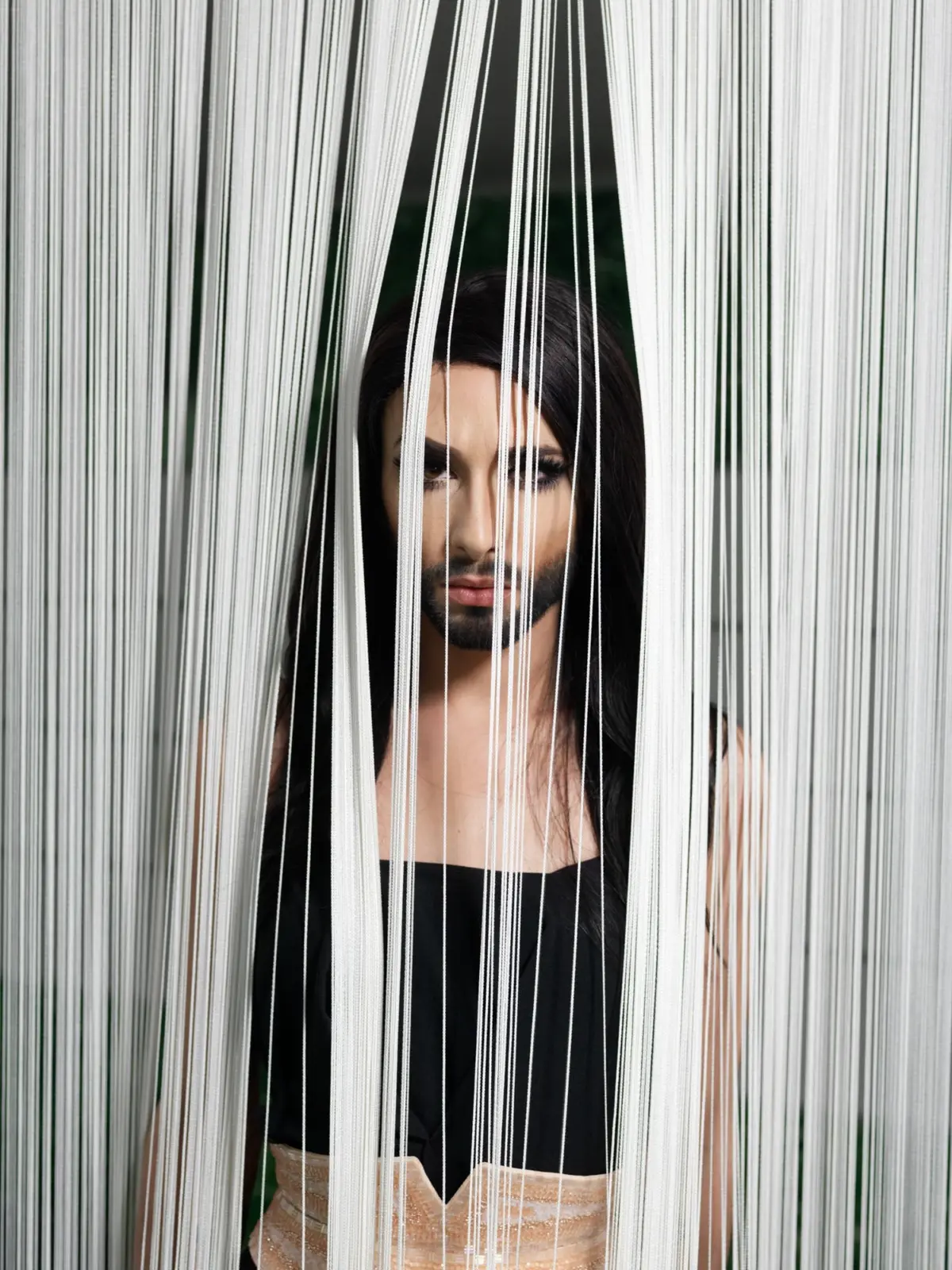 Person with long dark hair behind vertical white strings, wearing black top and peach skirt, peering through a string curtain outdoors.