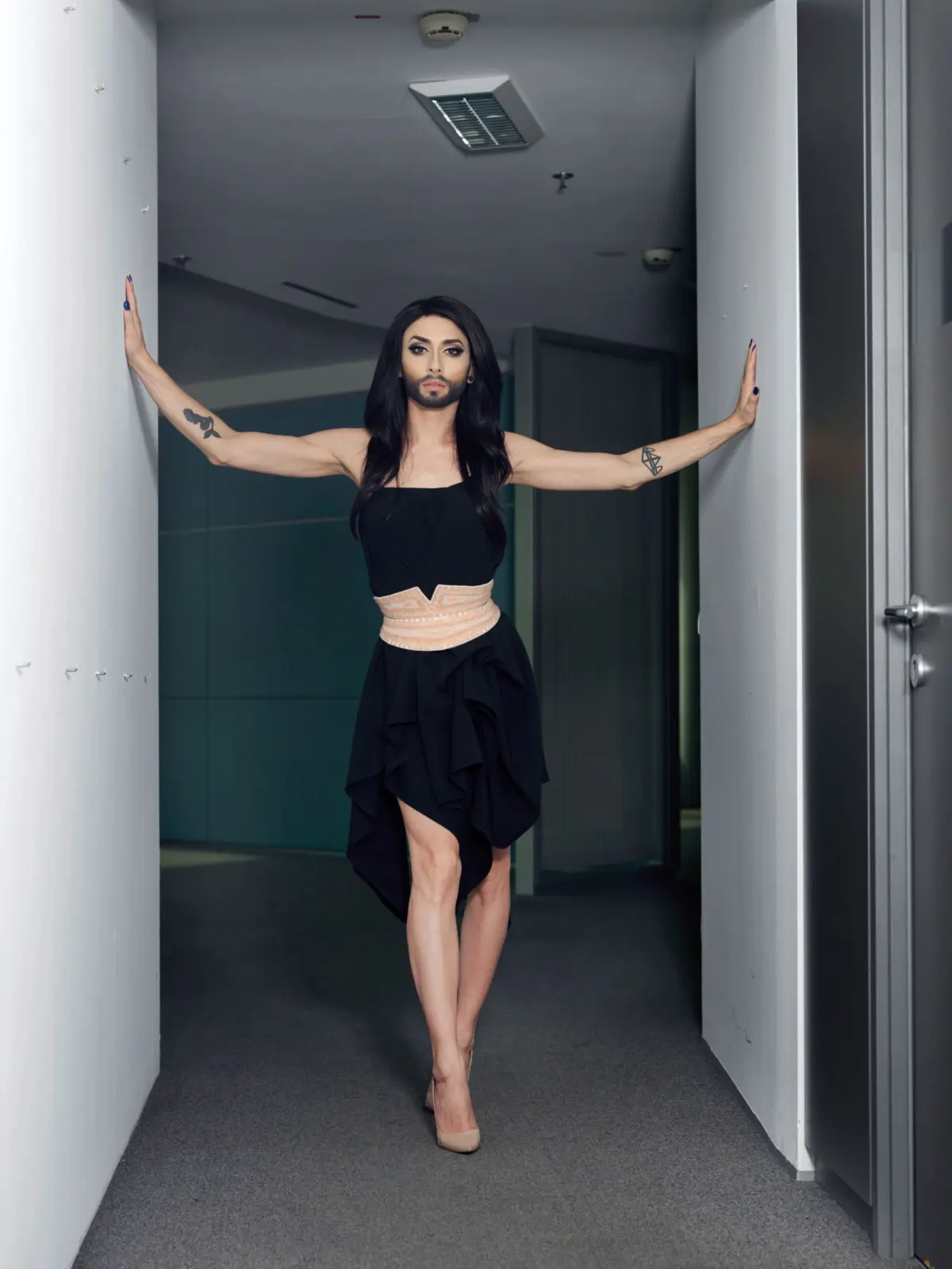 Person with long black hair and makeup wearing a black dress with beige waist belt stands in a narrow hallway, arms pressed to white walls.