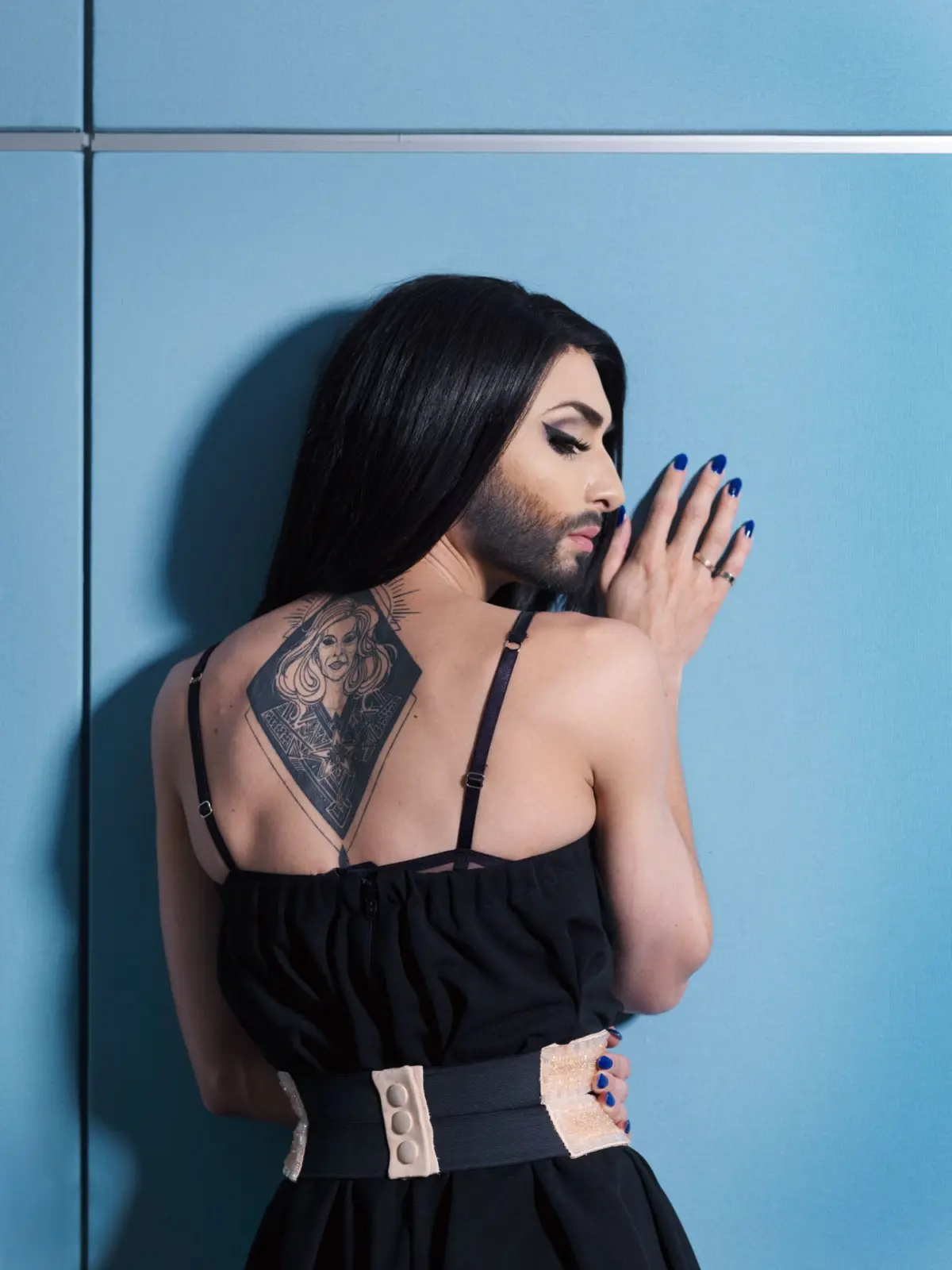 Person with long black hair leans against a blue wall, hands on wall; black sleeveless top, beige belt, geometric back tattoo visible.