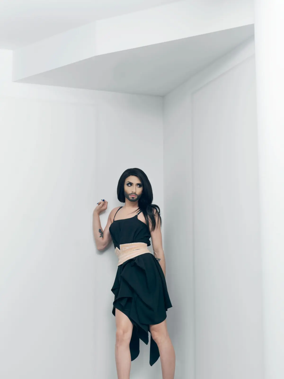 Person with long black hair and makeup wearing a black dress with beige waist belt, standing in a white corner of a minimalist room.