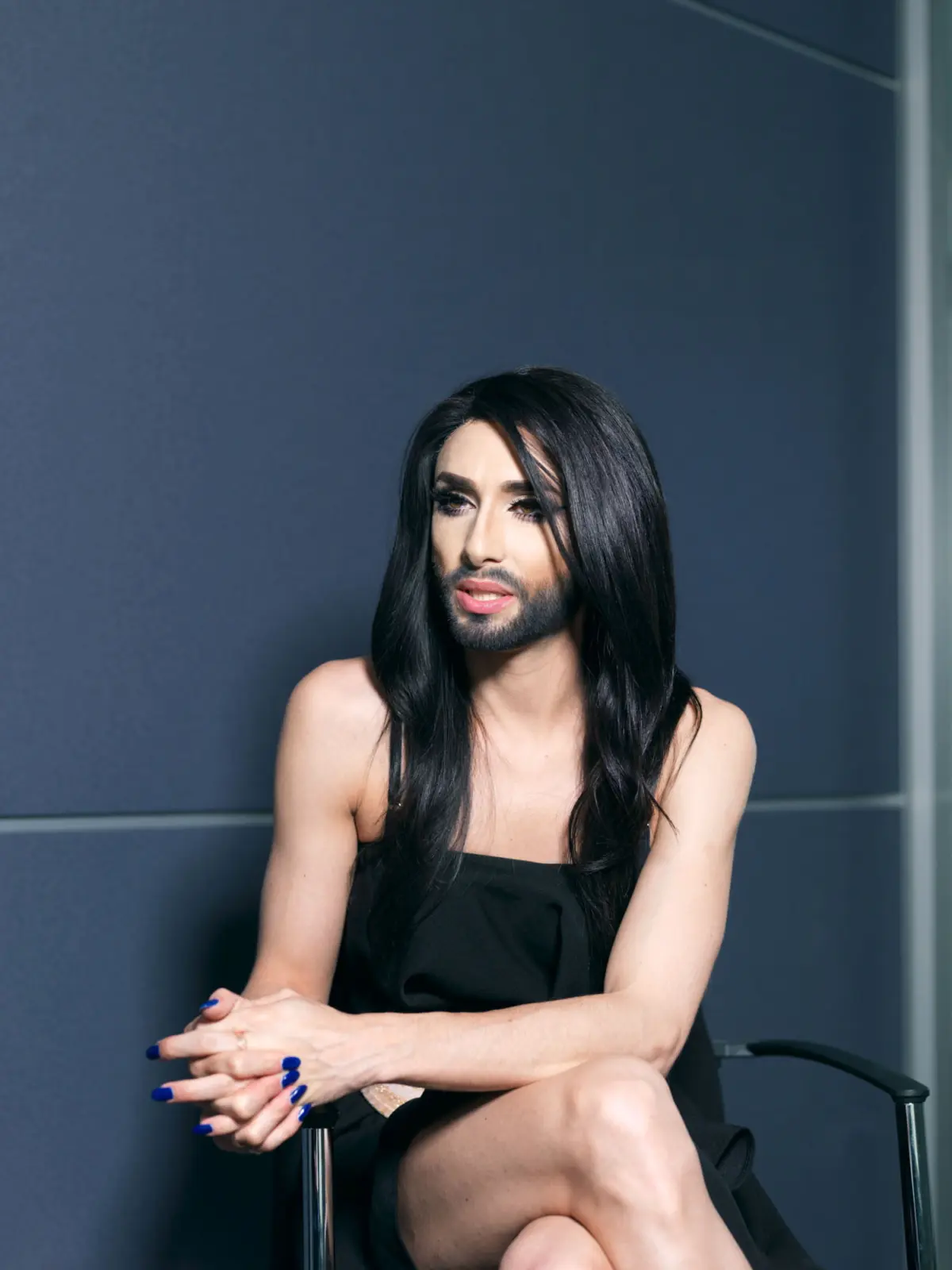 Person with long black hair, makeup, and a black dress sits on a metal chair with hands clasped in lap against a dark blue wall.