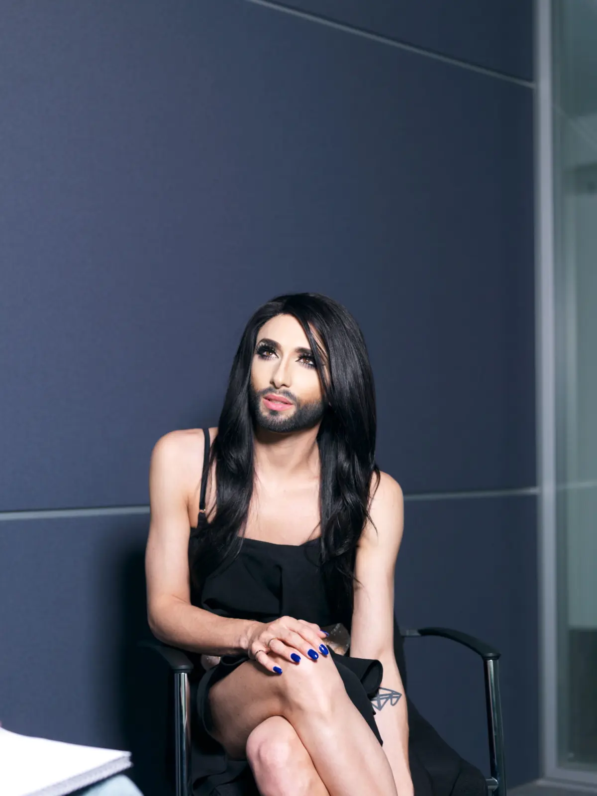 Person with long black hair and makeup sits in a black dress on a chair against a dark blue wall.