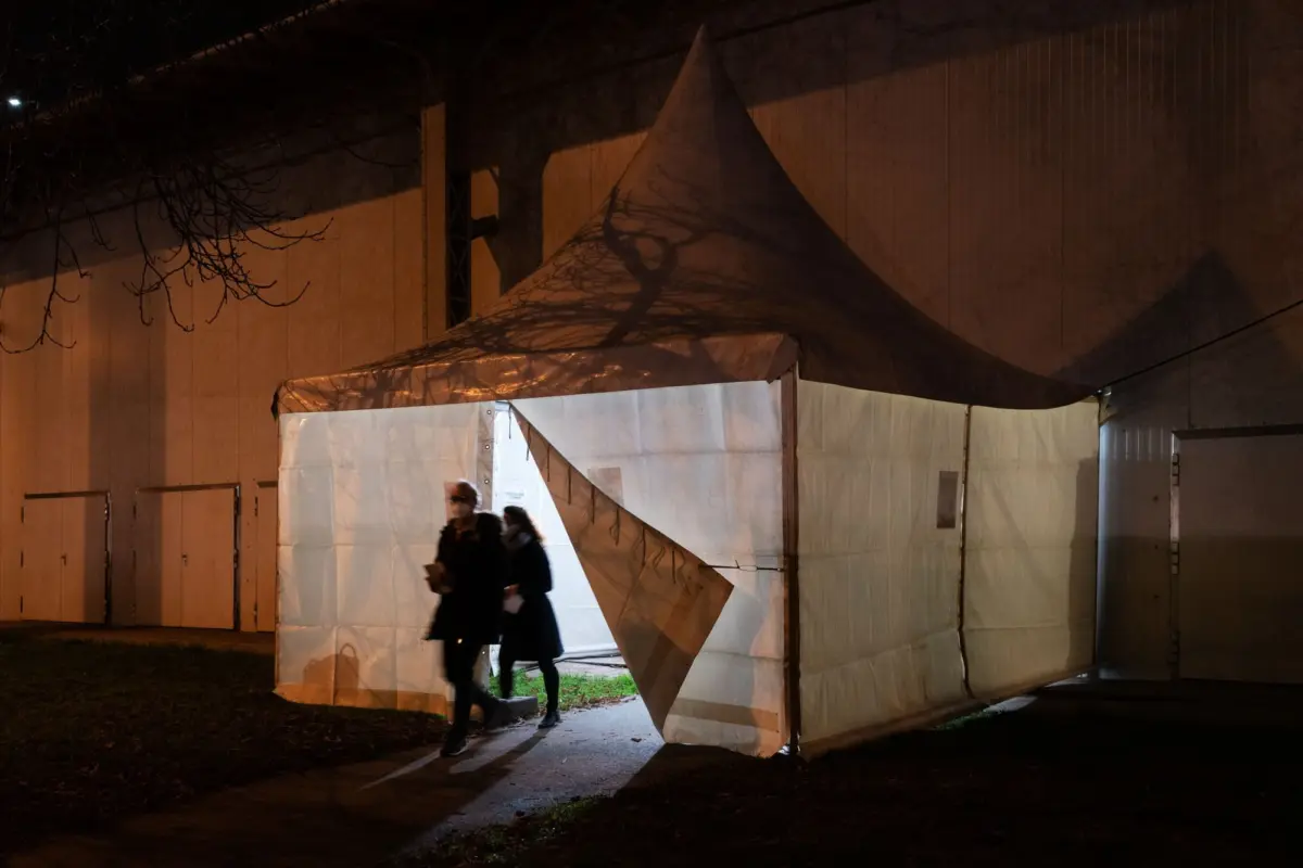Night scene of a white peaked-roof canopy tent beside a wall; two pedestrians walk past, with tree branches casting shadows on the tent.
