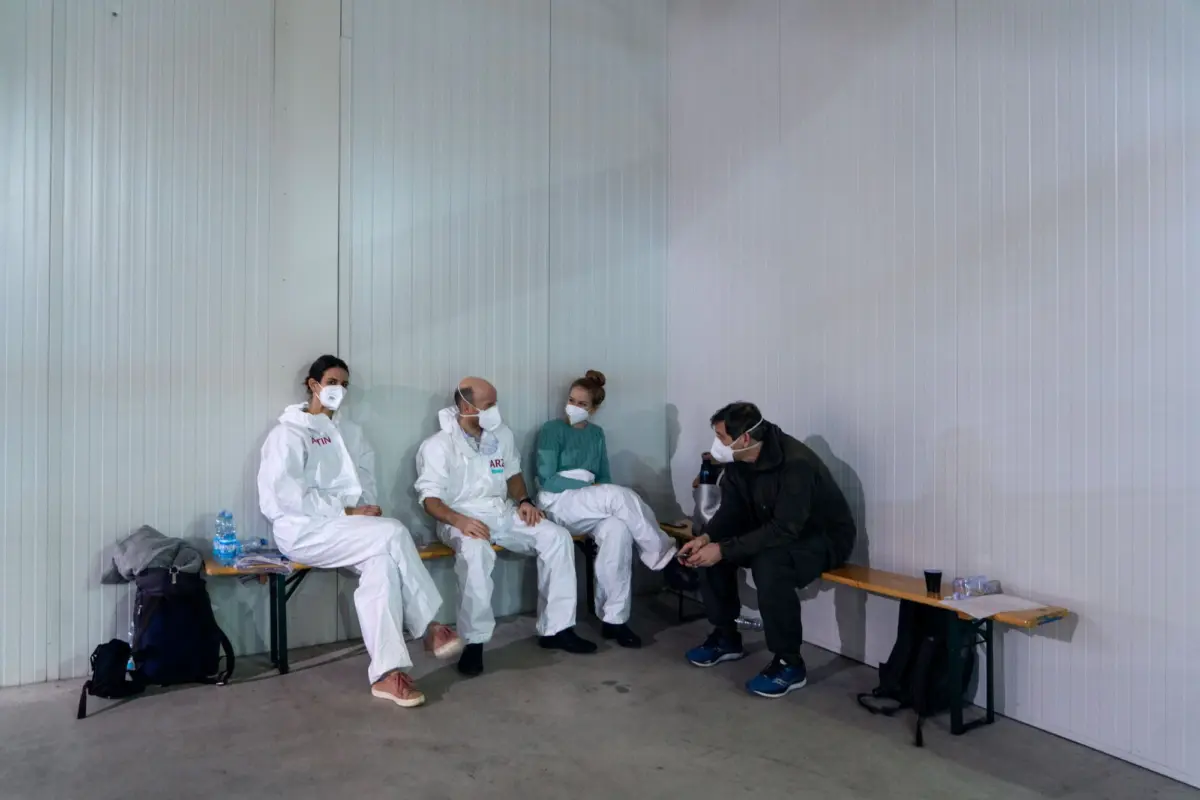 Four people in protective suits and masks sit on benches along a pale corrugated metal wall indoors, talking.