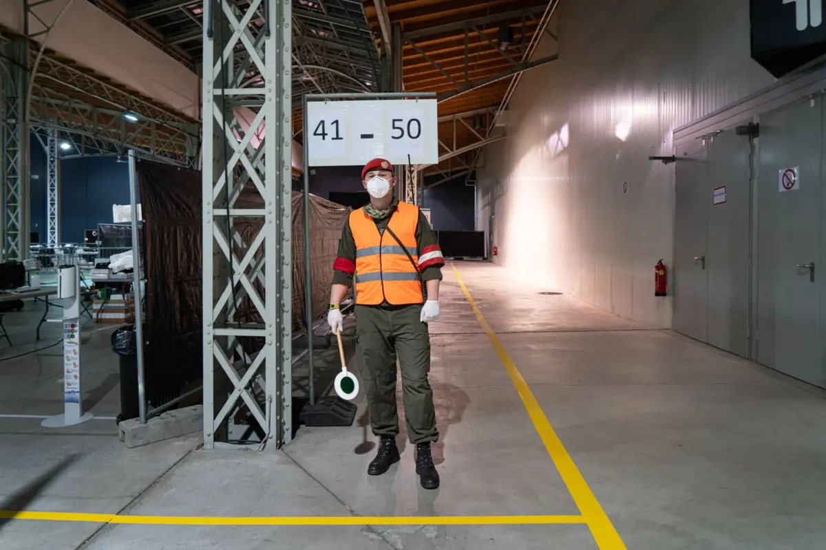 Uniformed security guard in orange safety vest and red beret, masked, holding a baton, beside a 41-50 sign in an industrial warehouse.