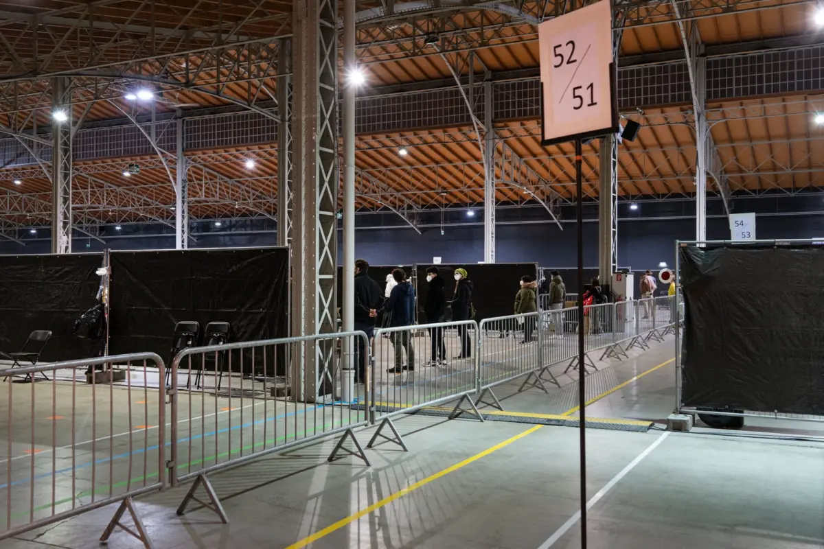 People queue behind metal barriers inside a large indoor exhibition hall with a truss ceiling; sign reads 52/51.