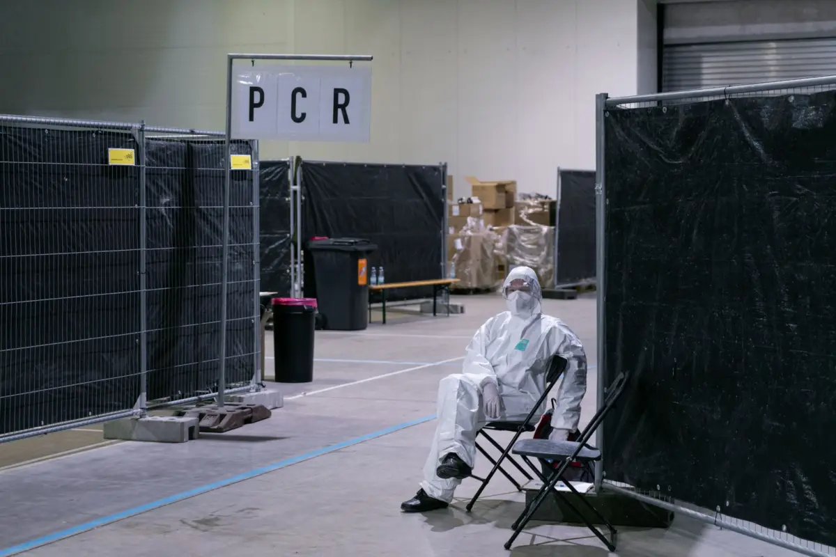 Person in white hazmat suit seated on folding chair inside a large testing area with a PCR sign and black privacy screens.