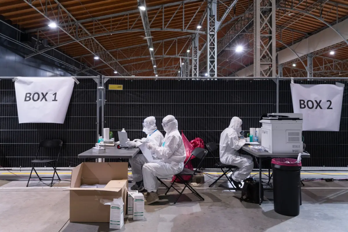 Healthcare workers in hazmat suits at desks processing tests in a large warehouse; banners read Box 1 and Box 2.
