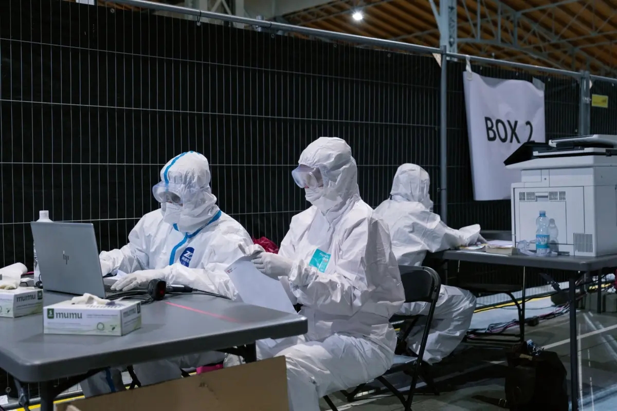 Hazmat-suited technicians at a temporary testing station, reviewing documents beside a laptop; "Box 2" banner in a fenced industrial setting.