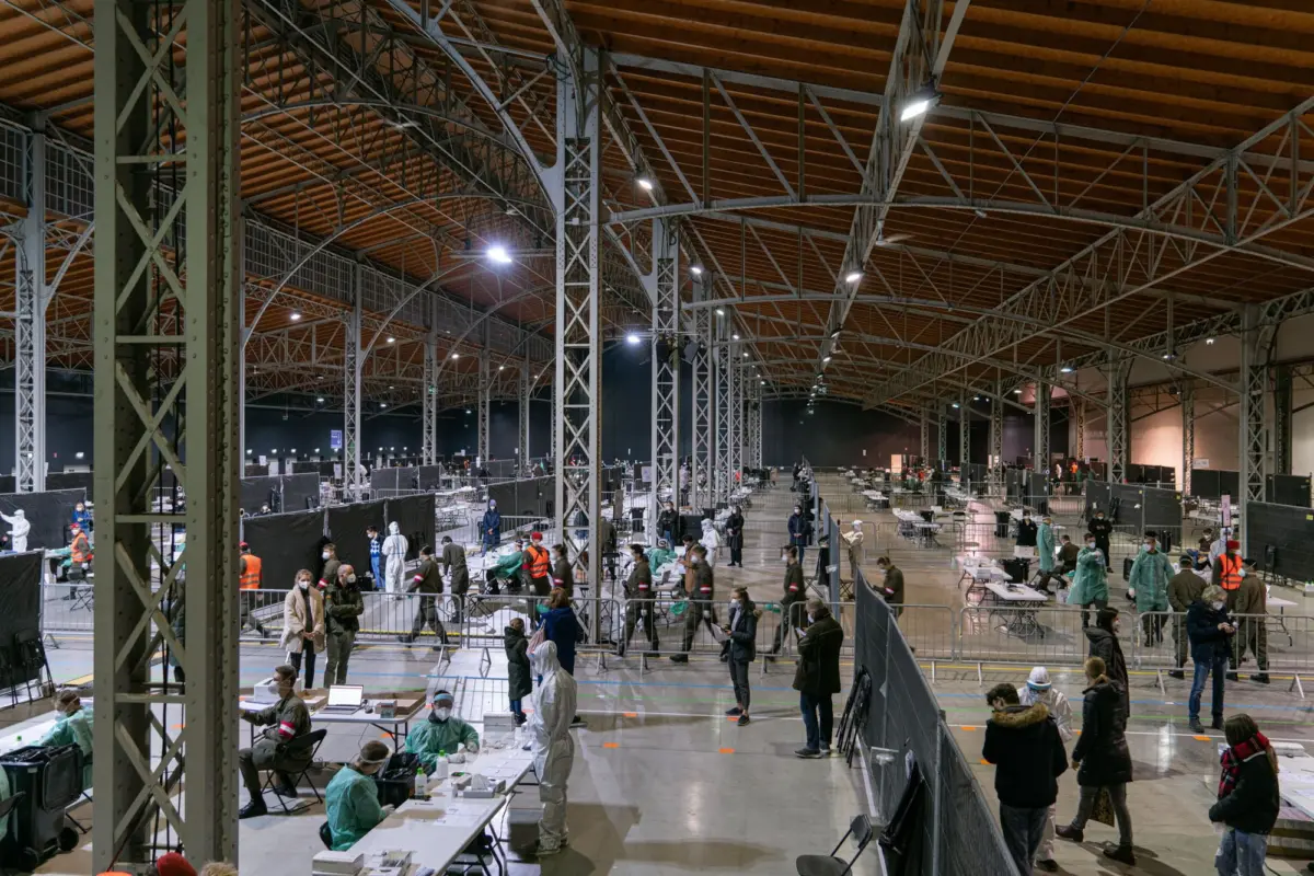 Warehouse-style hall with steel trusses; healthcare workers in PPE at desks assist people queuing for vaccination at a mass site.