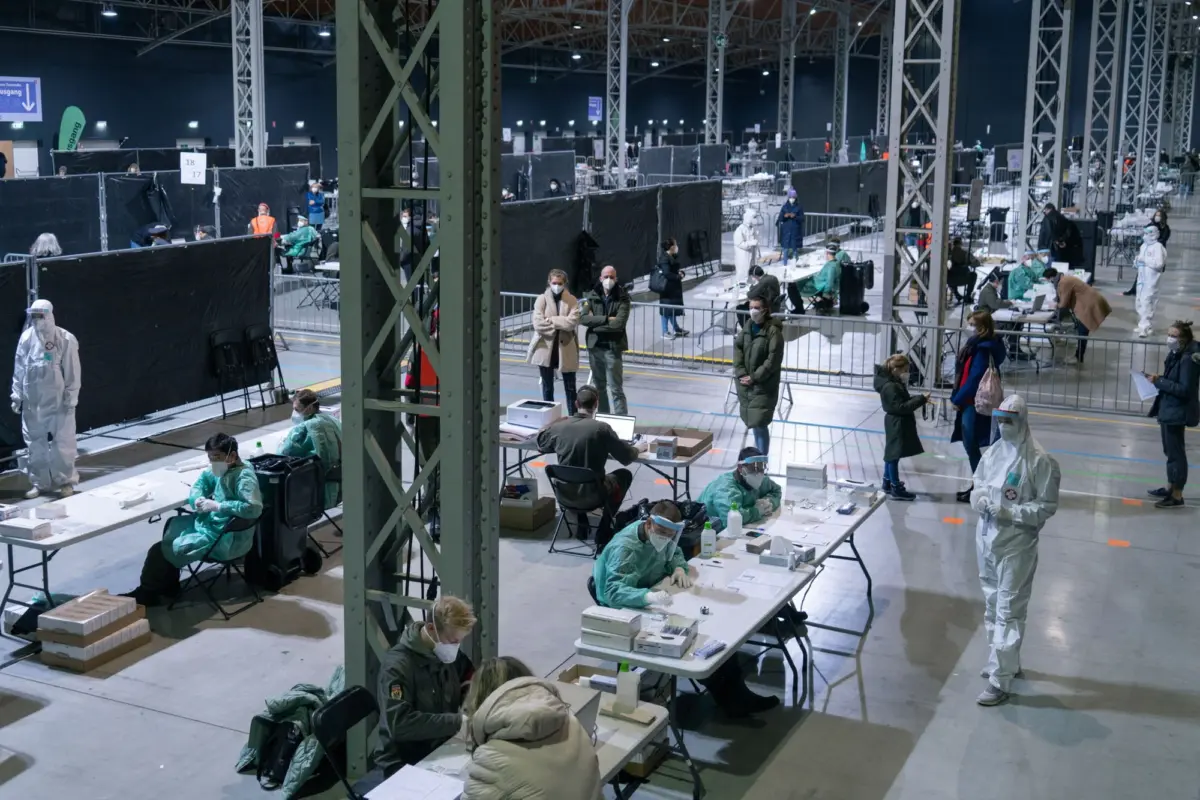 Large indoor vaccination center inside a hall with PPE-clad medical staff at desks and patients waiting behind black partition walls.