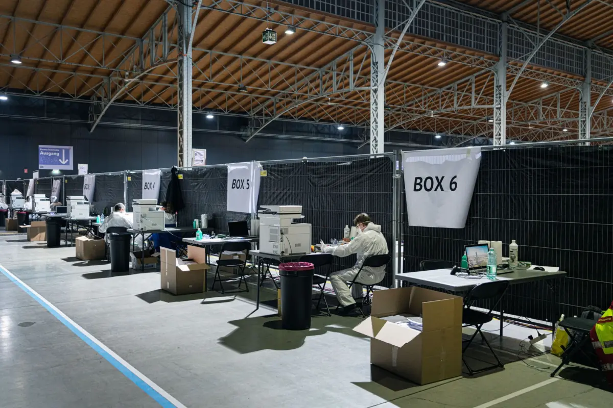 Healthcare workers in PPE at desks with printers inside a large hall; banners read BOX 4, BOX 5, BOX 6.