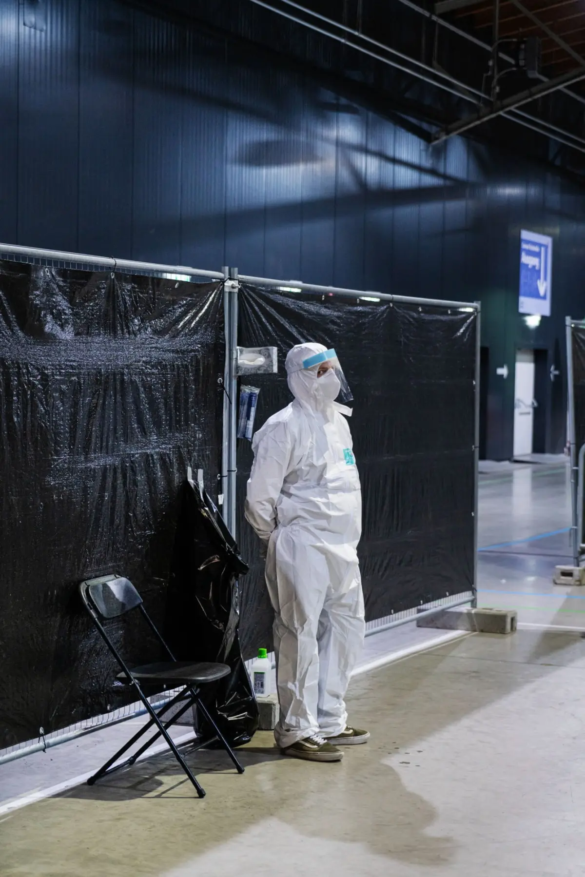 Hazmat-suited worker with face shield stands beside black plastic barrier partitions in an indoor testing area.