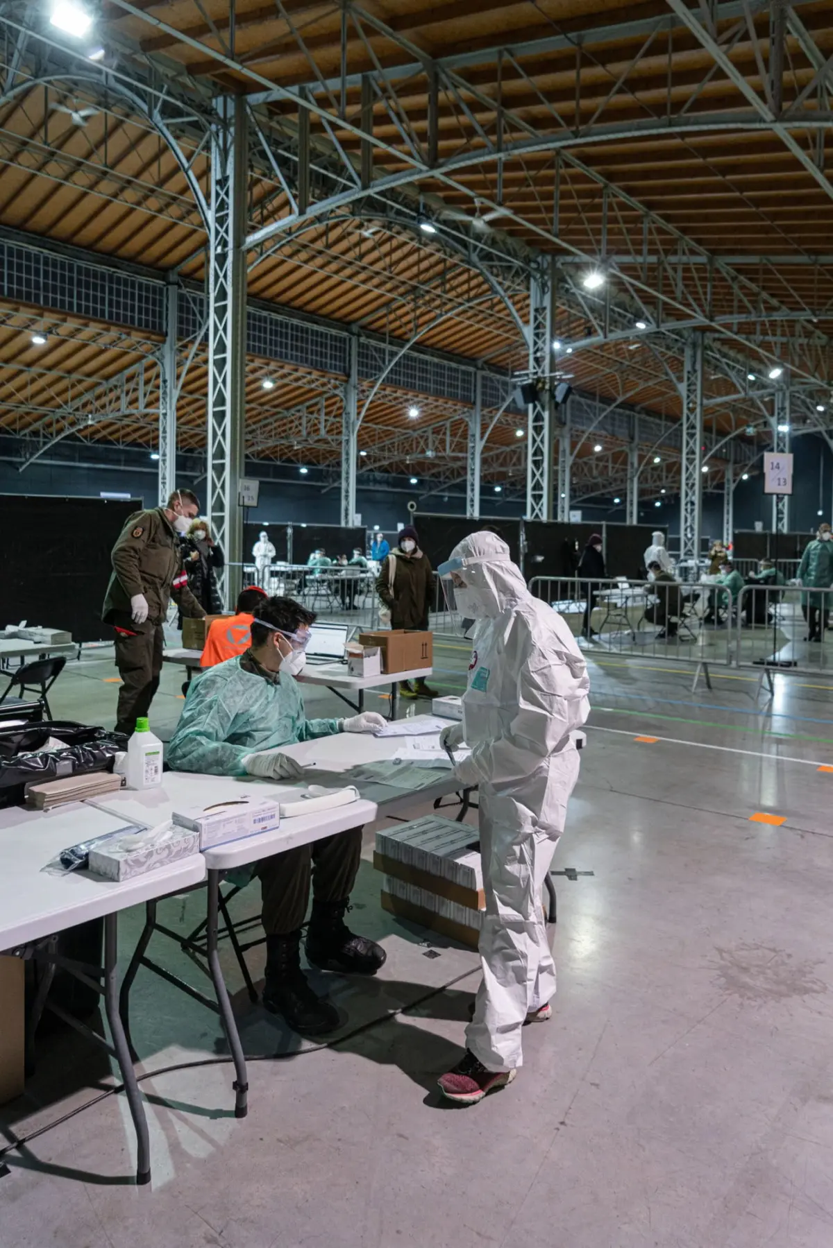 Person in white protective suit handles paperwork at a table in a large indoor medical processing center with steel structural beams.
