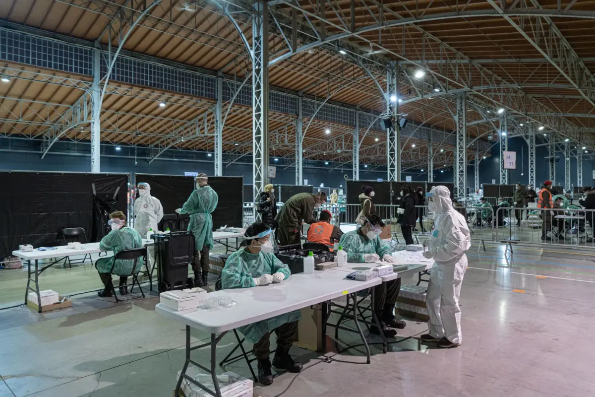 Medical staff in PPE vaccinating people at a large indoor vaccination center in a hall with metal trusses.