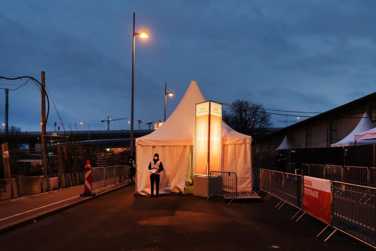 Evening outdoor Covid-19 testing site with white tents and orange 'CORONA TESTSTRASSE' signs; a staff member in a vest stands at the entrance.