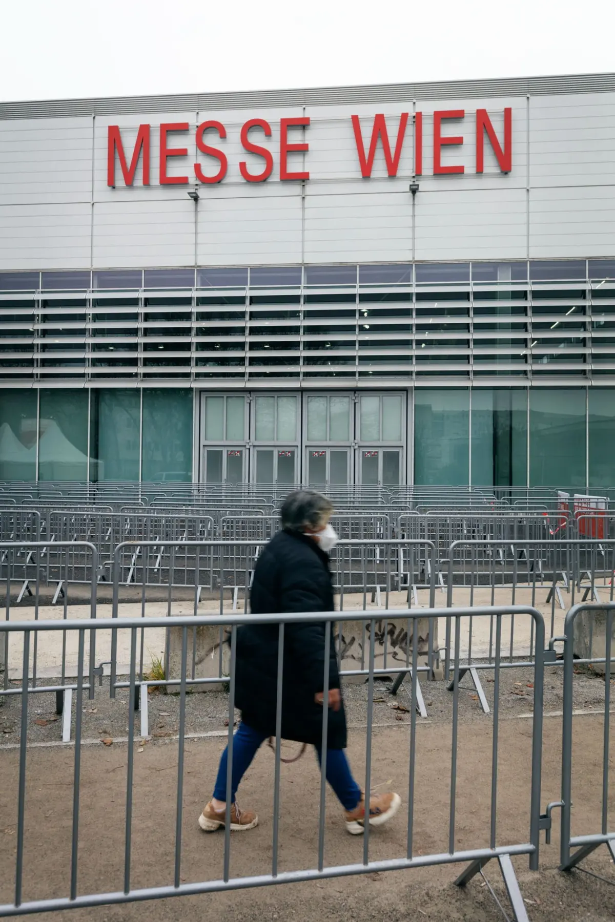 Person walking past metal crowd barriers outside the Messe Wien exhibition center; prominent red Messe Wien sign.
