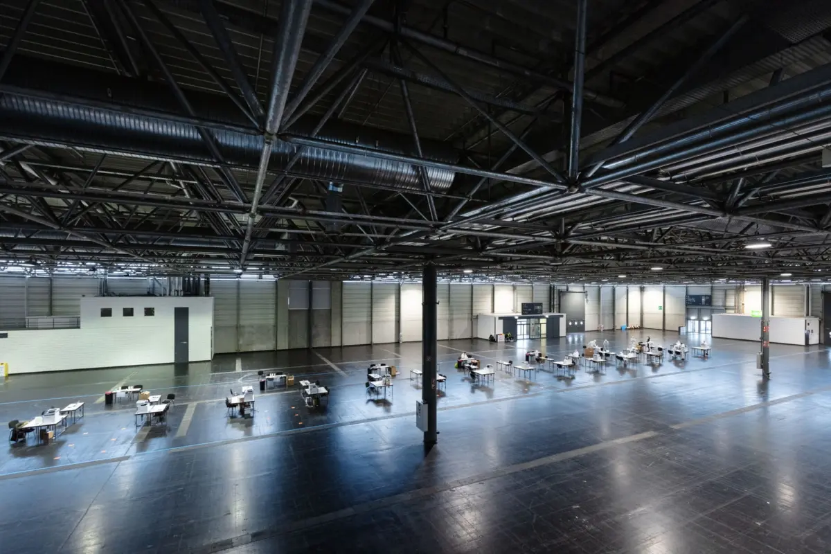 Interior of a large warehouse-like hall with a steel-truss ceiling; rows of tables and chairs arranged across the floor.