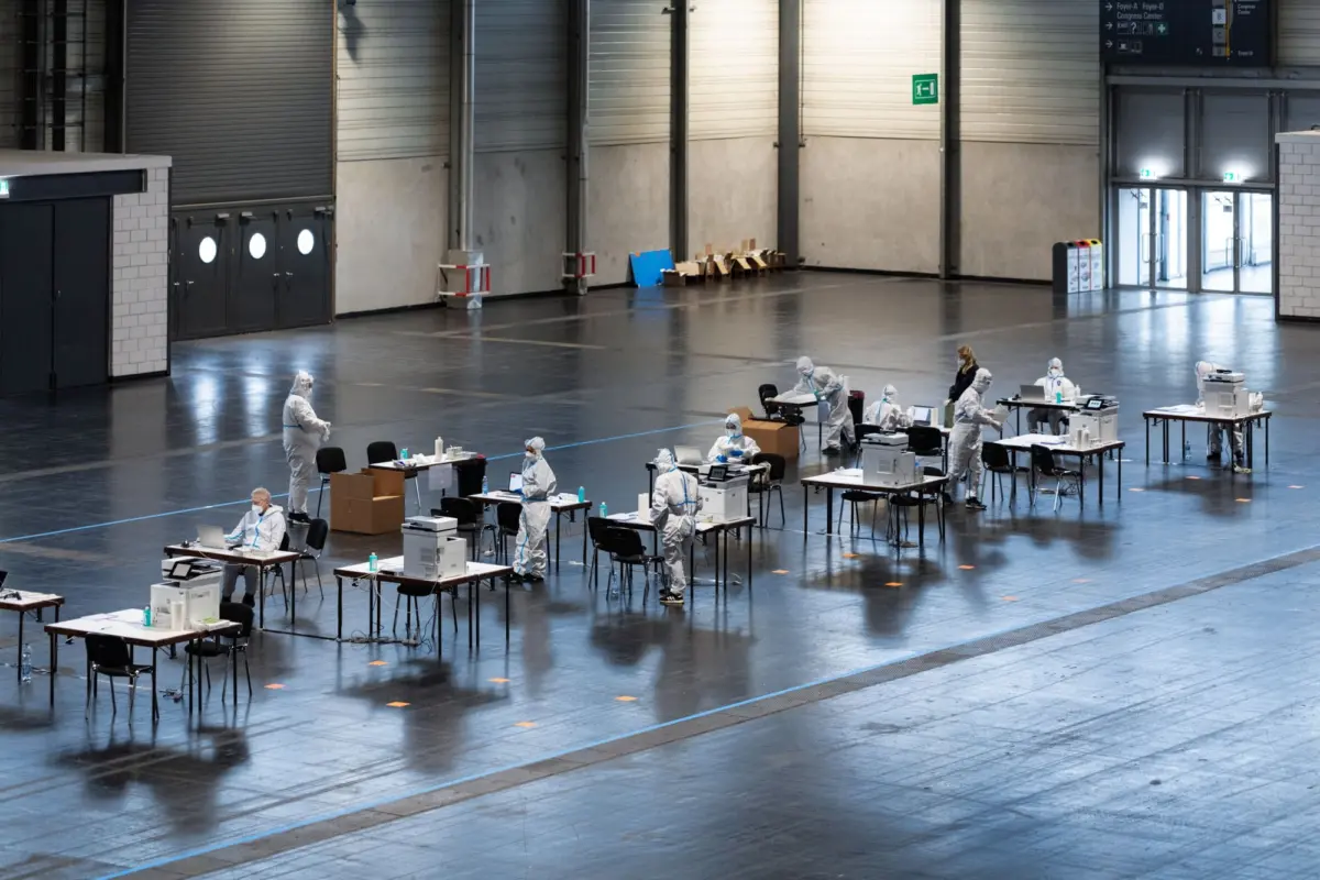 Workers in protective hazmat suits work at desks with laptops and medical supplies inside a large, empty warehouse.