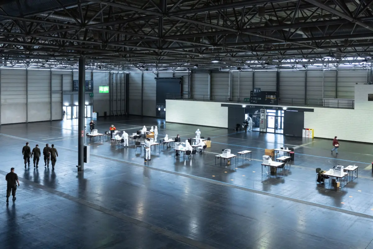Large warehouse hall with steel trusses; medical staff in protective suits at tables conducting testing.