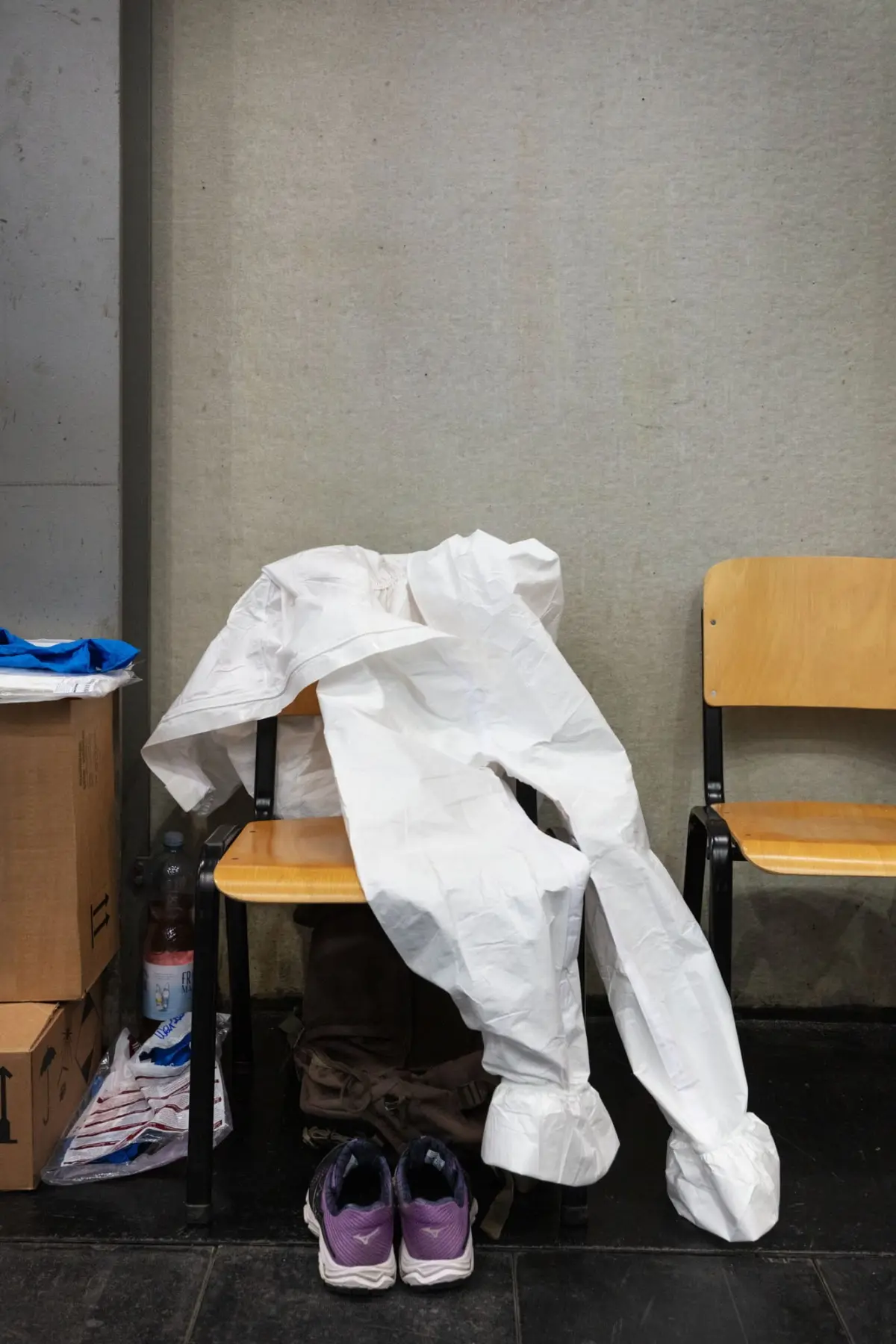 Crumpled white protective suit draped over a chair in a cluttered room with boxes, sneakers, and a bare concrete wall.
