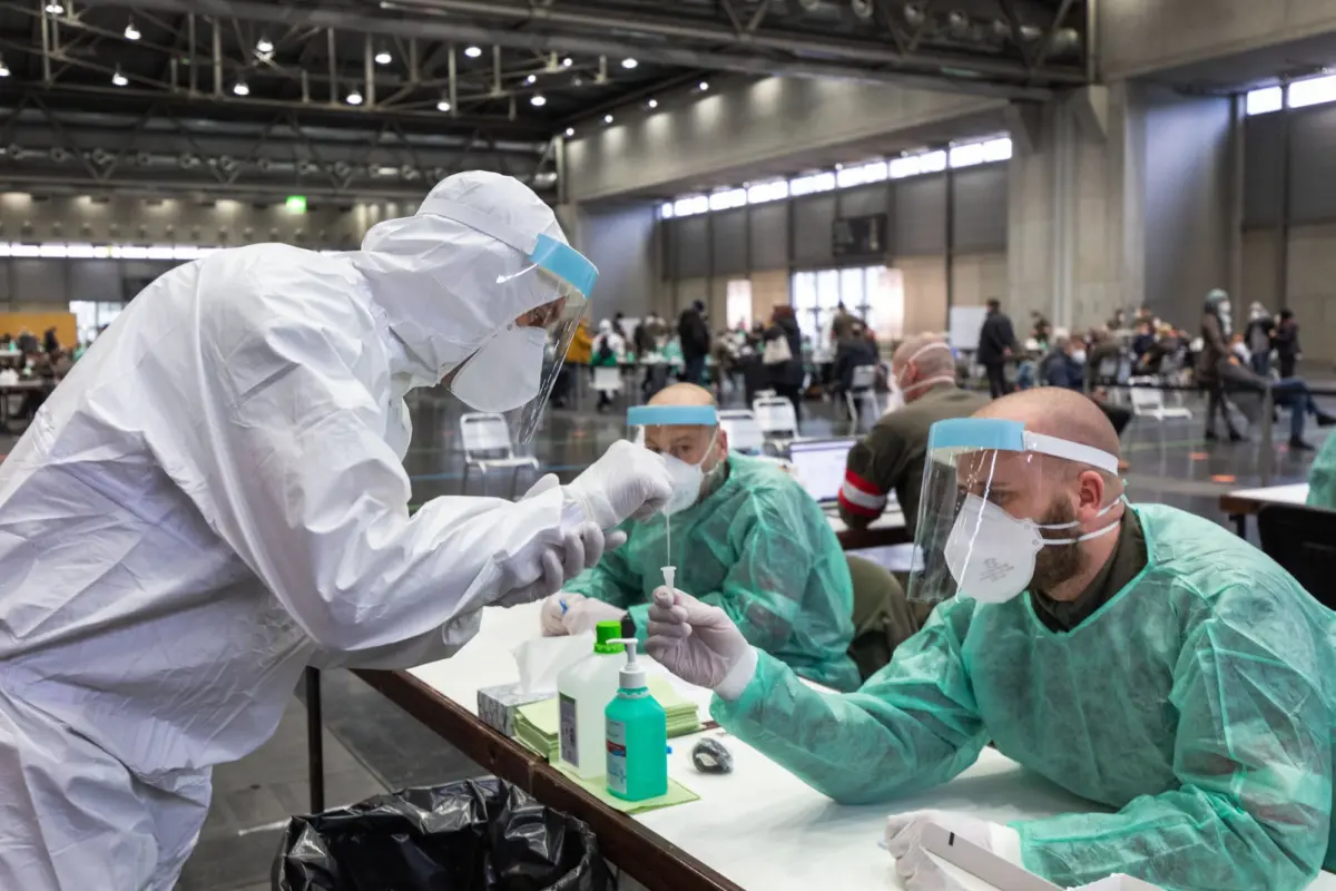 Healthcare worker in full-body protective suit and face shield administers vaccine to seated patient at a large indoor vaccination center.