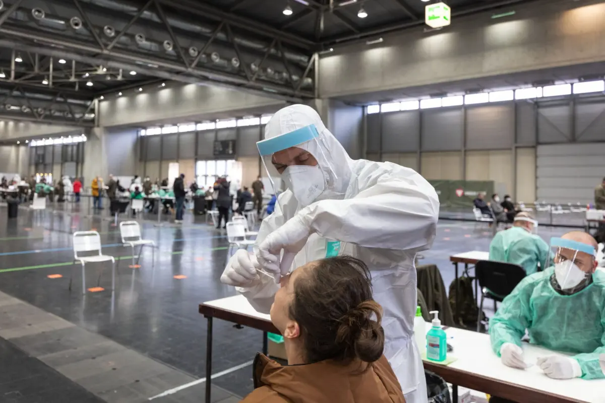 Healthcare worker in full PPE administers vaccine to seated patient at a large indoor vaccination center.