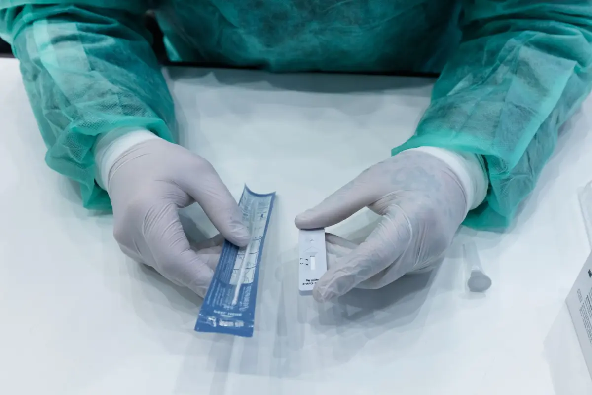 Medical professional in teal surgical gown and gloves handles a white rapid-test device with a blue strip on a sterile table.