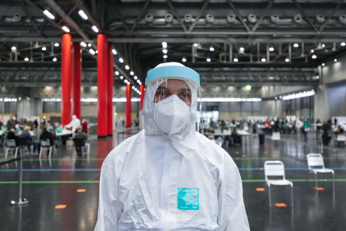 Healthcare worker in protective suit with face shield and mask stands in a large indoor vaccination center with red columns and seated people.