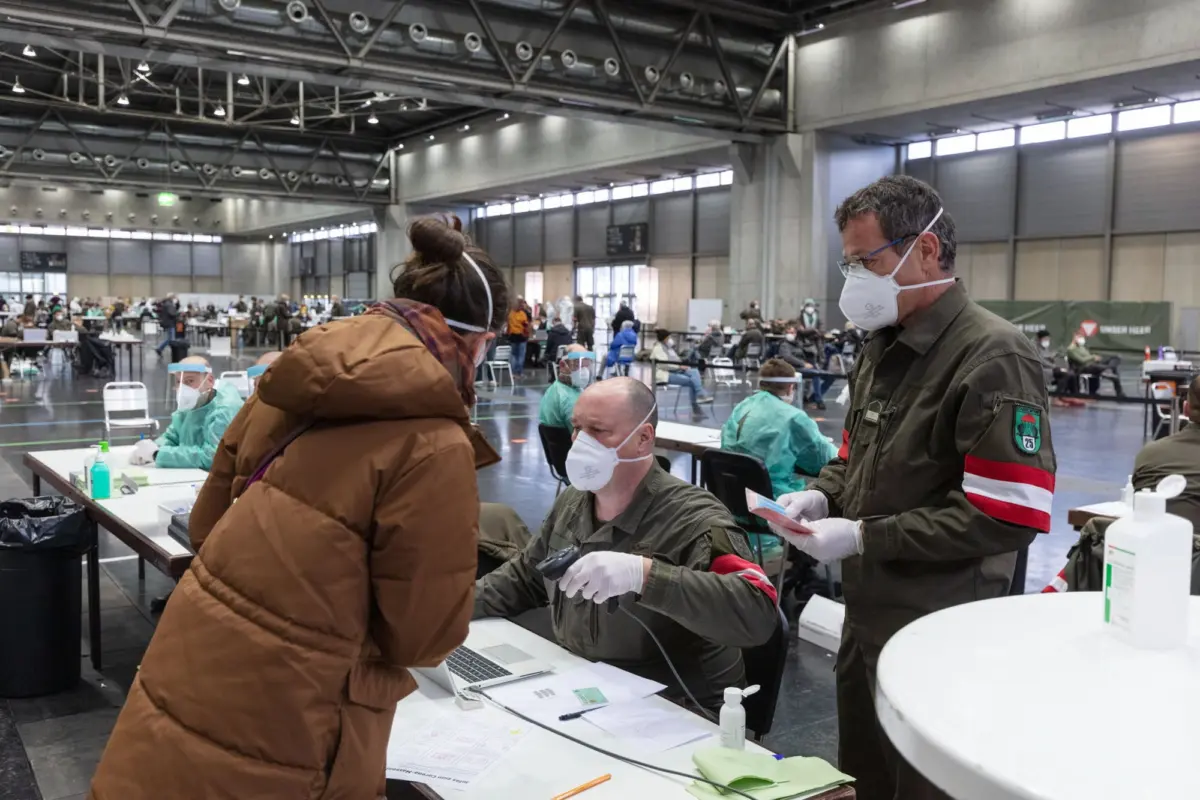 Healthcare workers in protective gear vaccinate a seated patient at a large indoor vaccination center.
