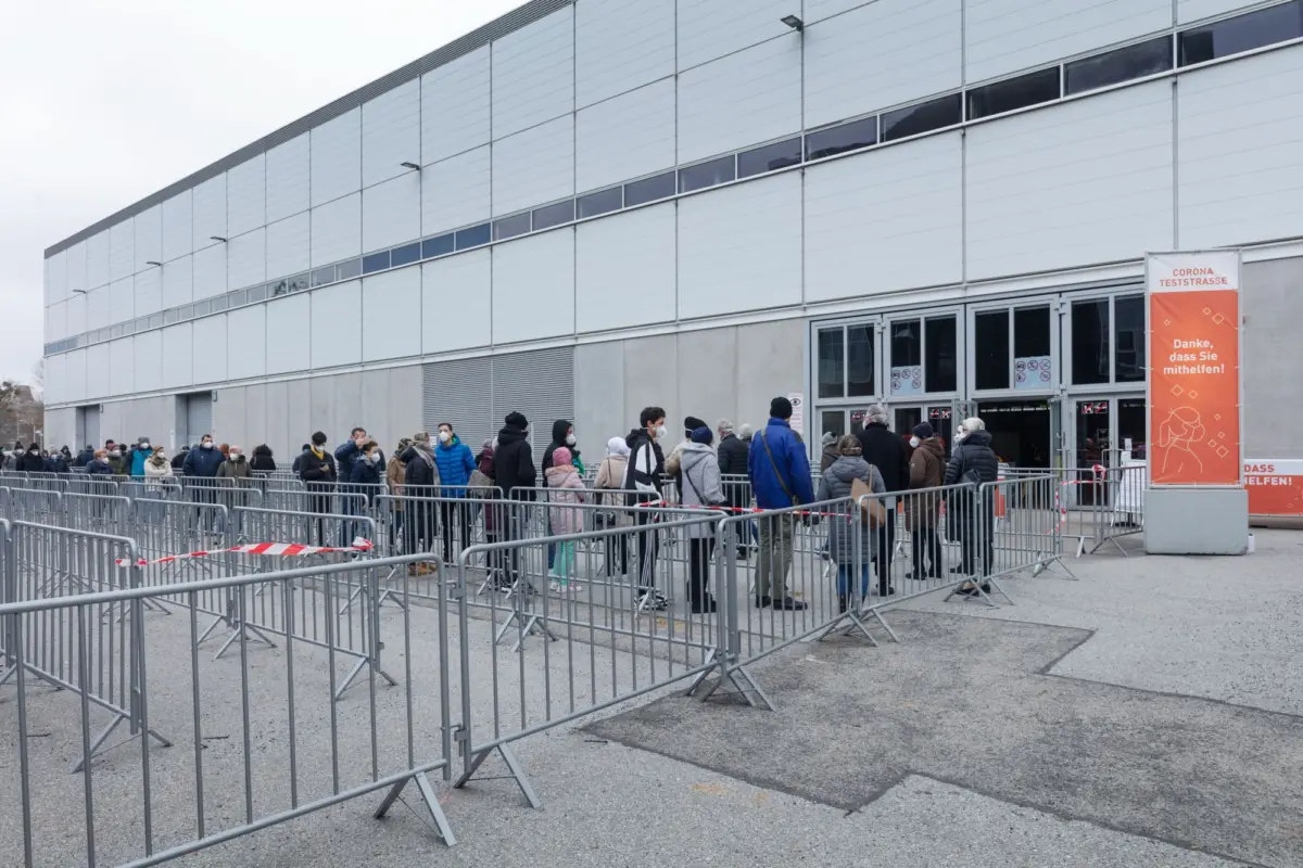 Queue of people wearing masks outside a modern grey building; prominent orange Corona Teststrasse sign at entrance.