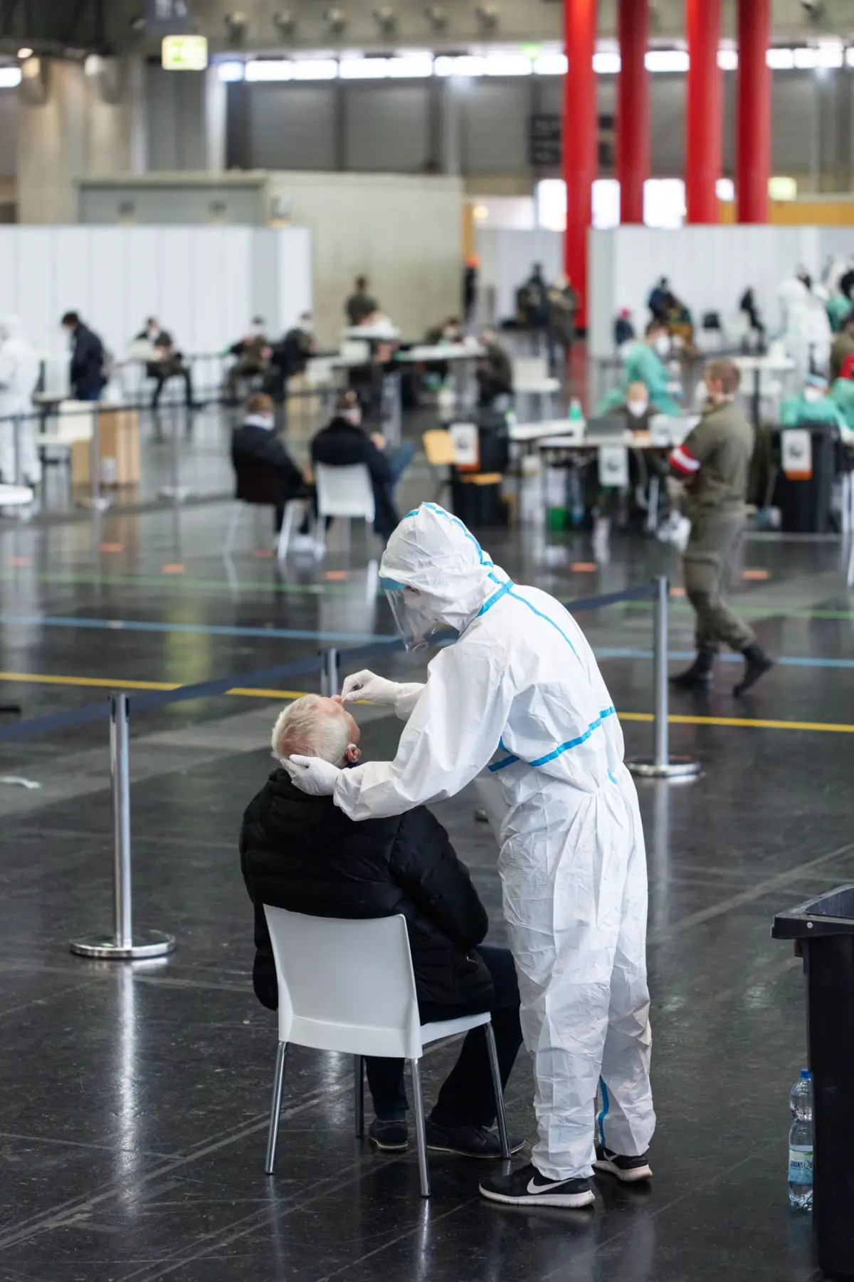 Healthcare worker in full PPE administers a vaccination to an elderly person at a large indoor vaccination center.