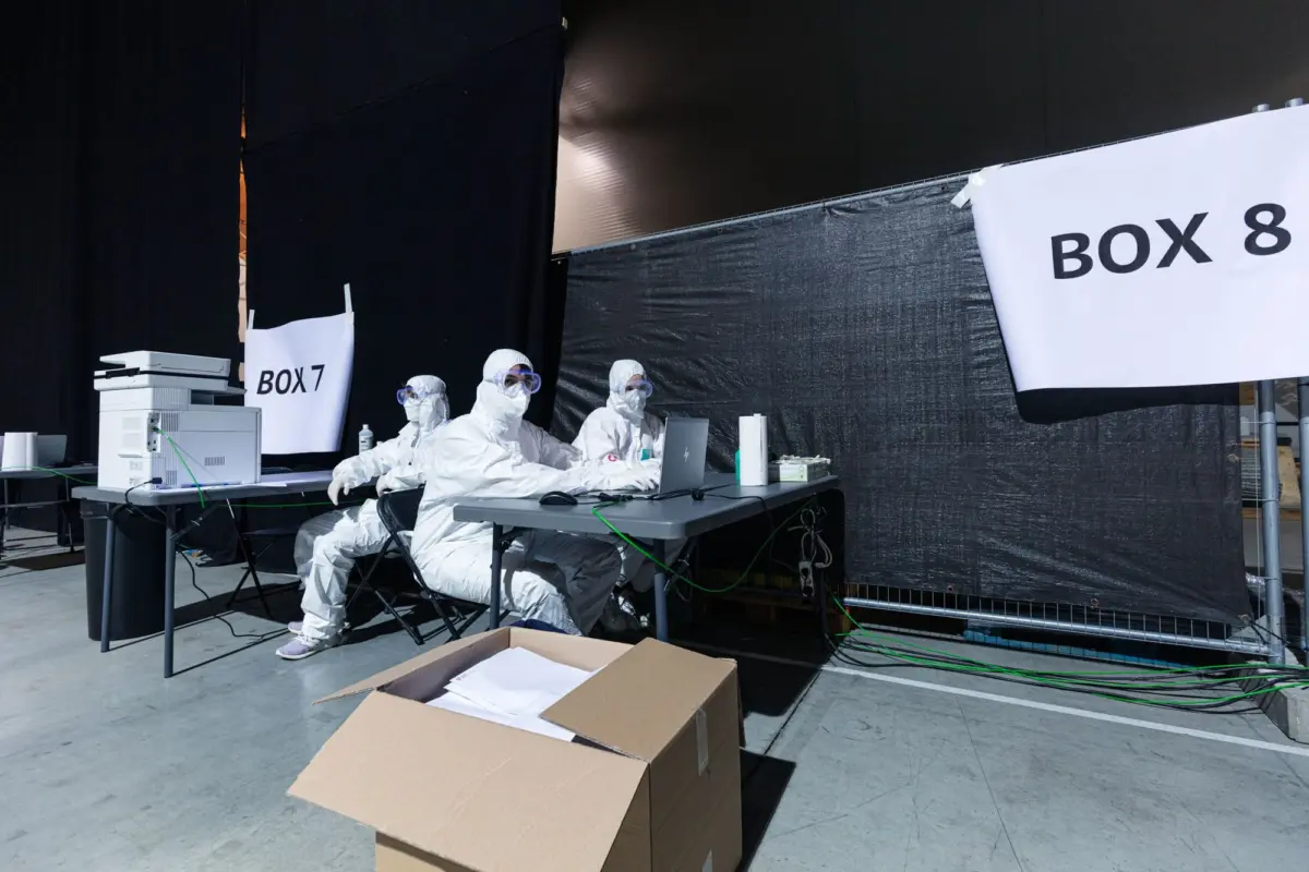 Hazmat-suited technicians work at a table with laptops and lab equipment inside a black temporary lab; signs read BOX 7 and BOX 8